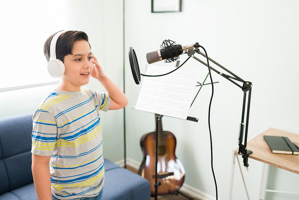 A boy wearing headphones stands in front of a microphone, singing or speaking, with a music stand and guitar in the background—perfect for those searching for singing classes for kids near me.
