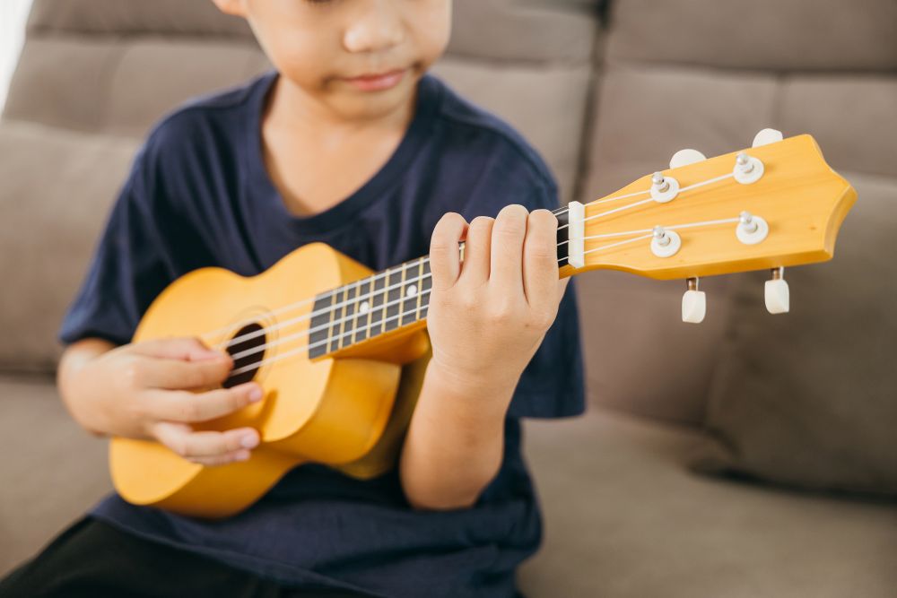 A child sitting on a couch plays a small yellow ukulele, focusing on pressing the strings with one hand and strumming with the other—perfectly capturing the joy of ukulele lessons for kids Singapore offers.