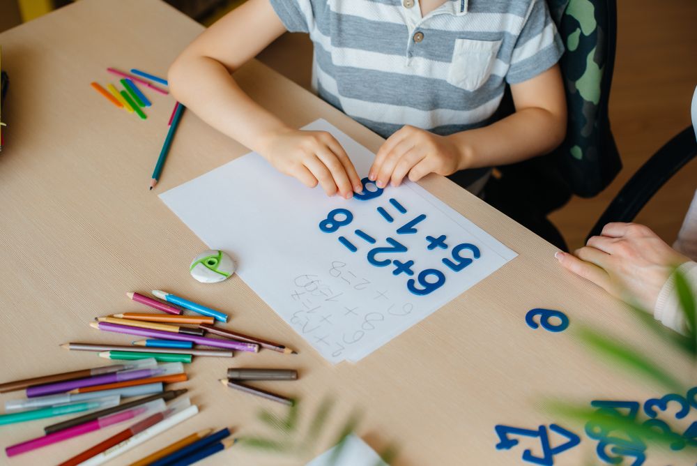Child sitting at a desk solving simple math equations with blue plastic numbers and colored pencils scattered around, exploring concepts from Math Sec 1: A Guide for Parents and Students.